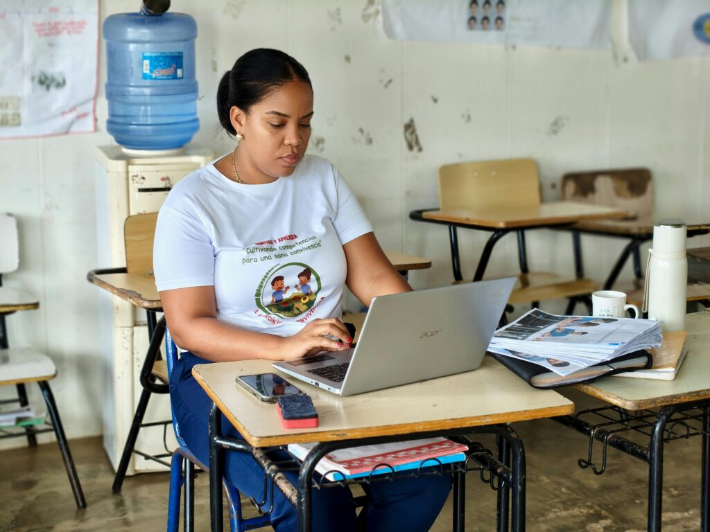 Adult woman typing on a laptop in a classroom, surrounded by desks and textbooks.